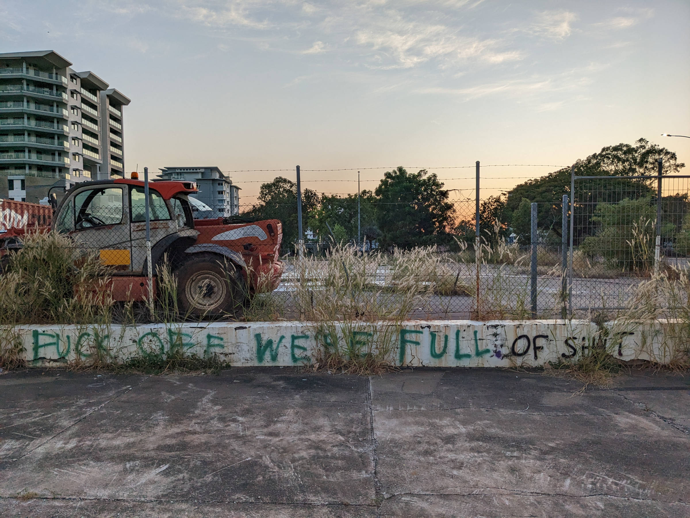 Photo of an overgrown parking lot in Darwin CBD at sunset, with a rusted out farm vehicle in the only occupied space. Graffiti has been sprayed crudely in black along the central wall, reads: "FUCK OFF WE'RE FULL". Somebody has added in green: "OF SHIT".